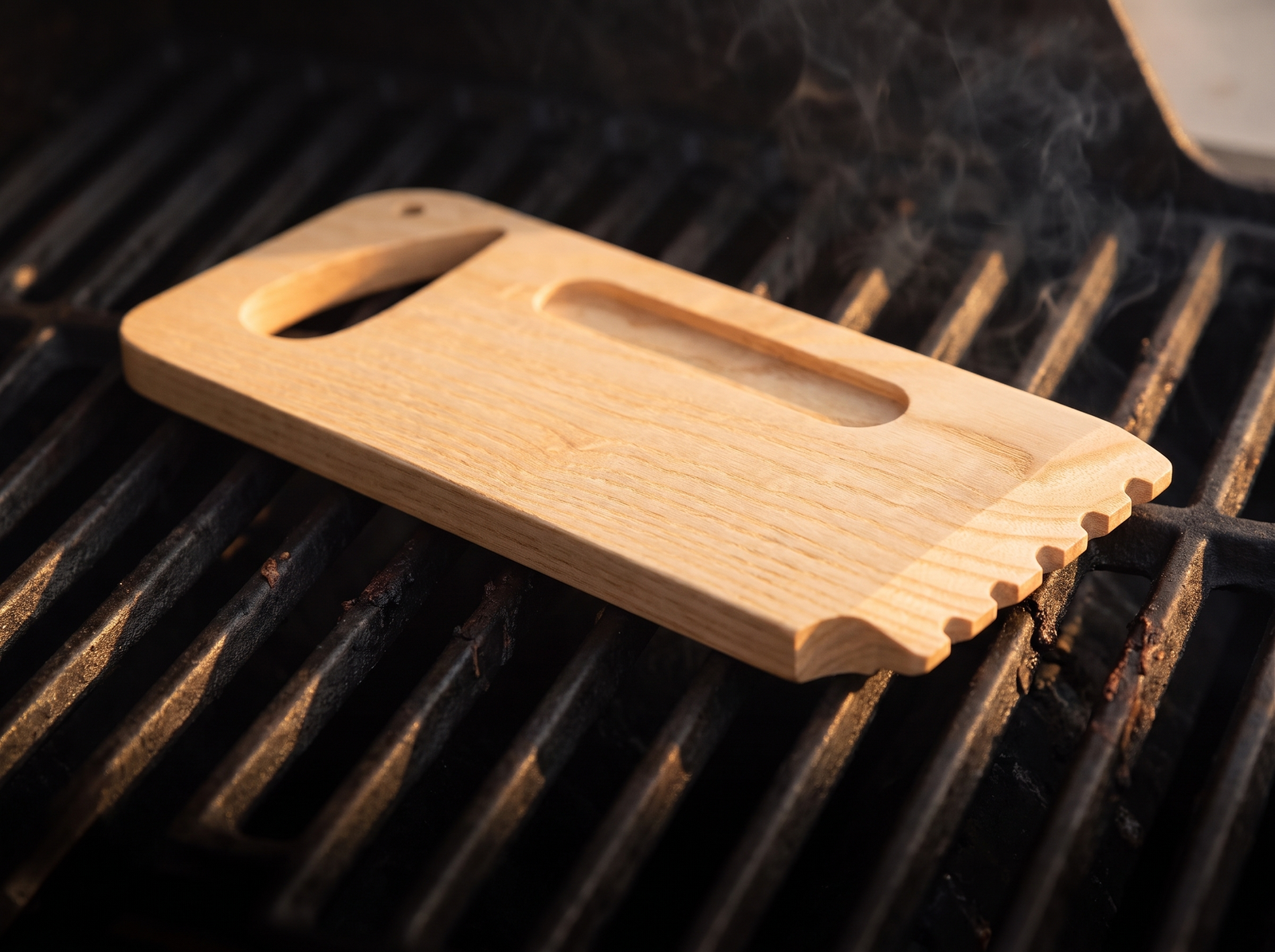 Solid ash wood grill scraper resting on cast iron grates in golden hour light