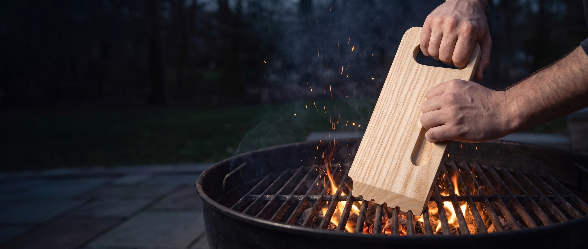Two hands scraping a hot charcoal grill grate with the Skrapa wooden scraper, full serrated edge on the bars, embers glowing below